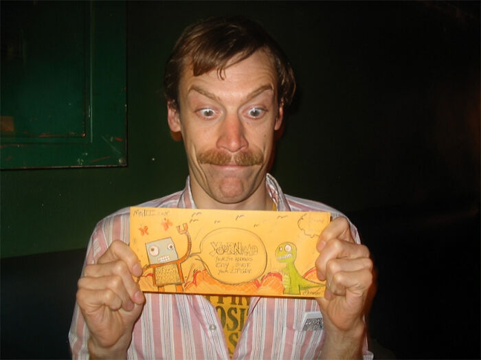 Man with mustache holding a colorful envelope, illustrating some realities of being a postal worker in a casual setting.
