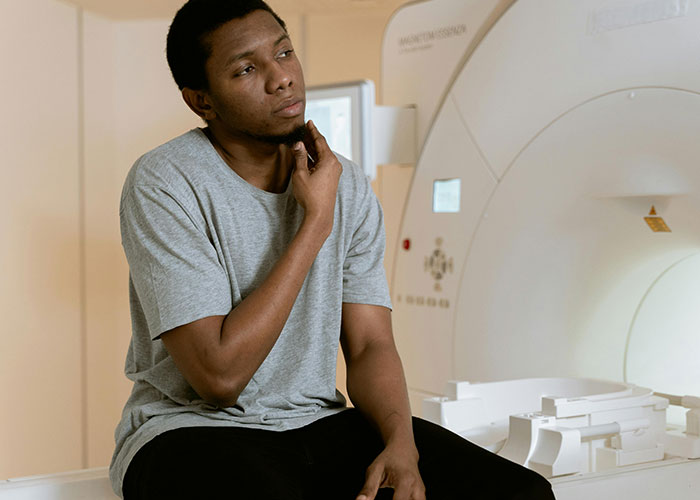 Man in a gray shirt looking thoughtful while sitting near a large medical imaging machine in a clinical setting.