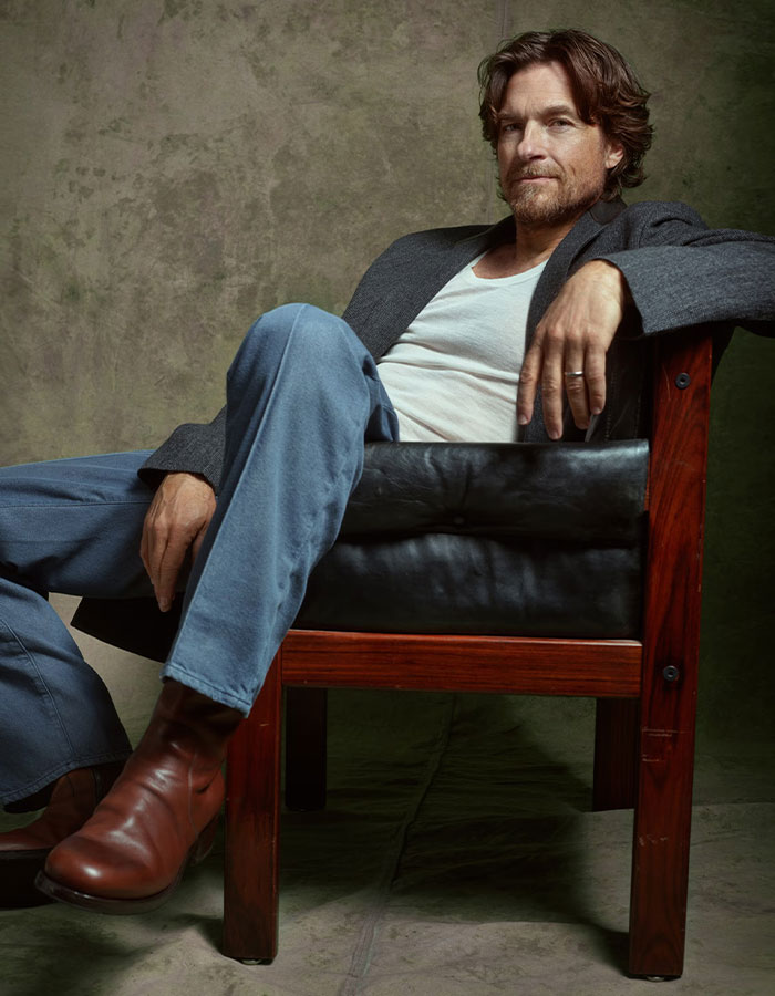 Jason Bateman sitting relaxed in a chair, wearing a gray blazer and brown boots, raising eyebrows with a remark about sister Justine