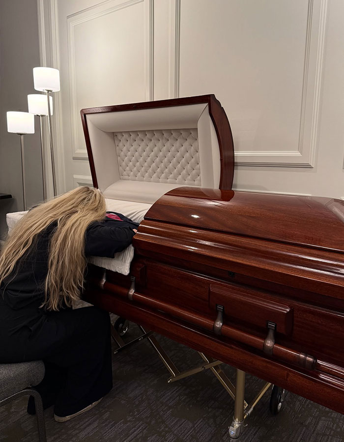 Woman with long blonde hair leans on a polished wooden casket in a subdued room, reflecting on Erika Kirk's curated moment.