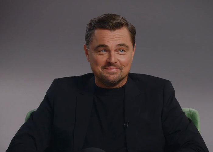 Leonardo DiCaprio in a black blazer sitting and smiling during an interview discussing his dating history.