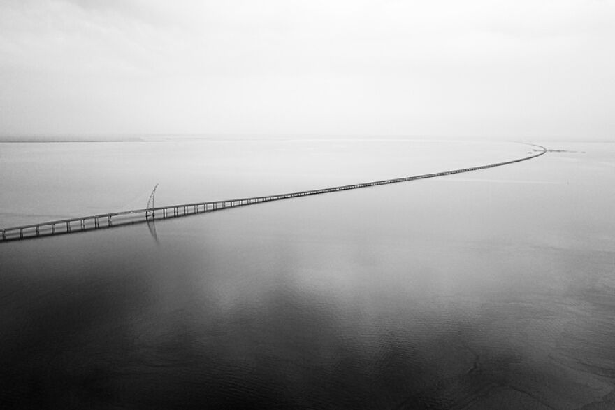 Aerial view of a remarkably engineered, extremely long bridge extending across calm water under a misty sky.