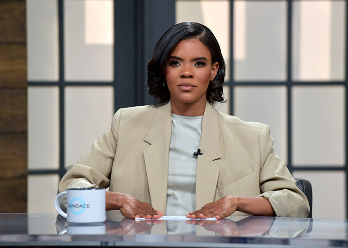 Candace Owens seated at a studio desk, wearing a beige blazer, discussing comments about Brigitte Macron.