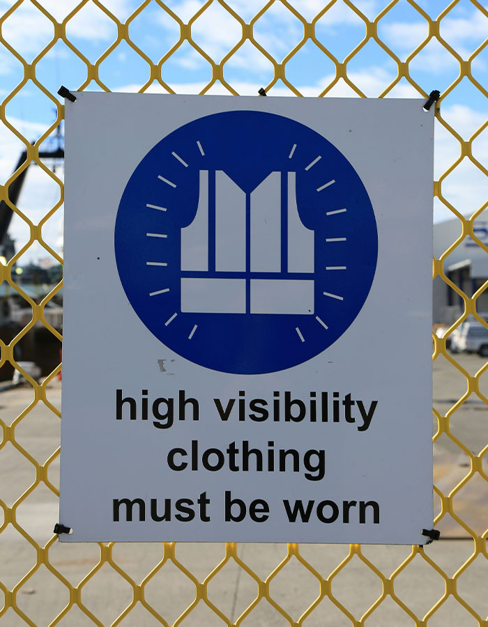Safety sign on yellow fence showing high visibility clothing must be worn to prevent forklift accident risks at worksite.