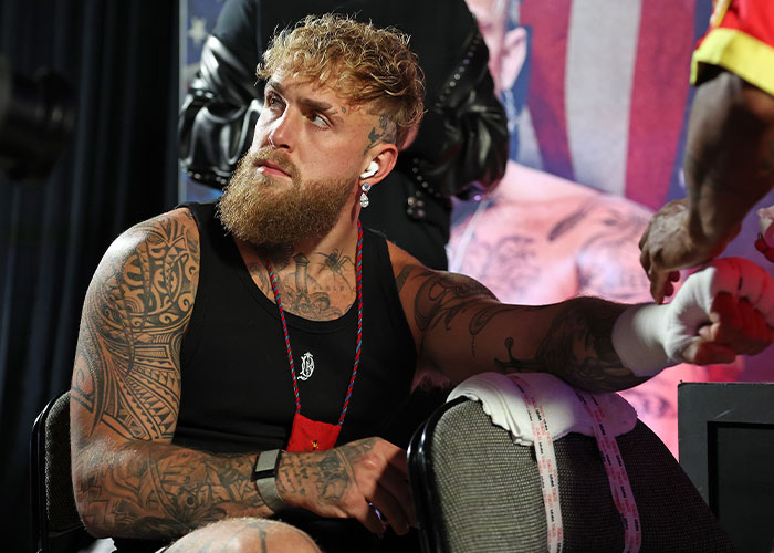 Jake Paul with tattoos and beard sitting wearing a black tank top, arm in a cast after broken jaw injury.