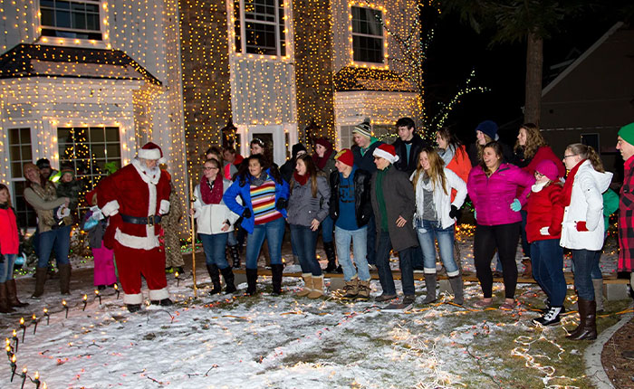 Group of people with a man dressed as Christmas lawyer celebrating outside a house decorated with holiday lights during winter.