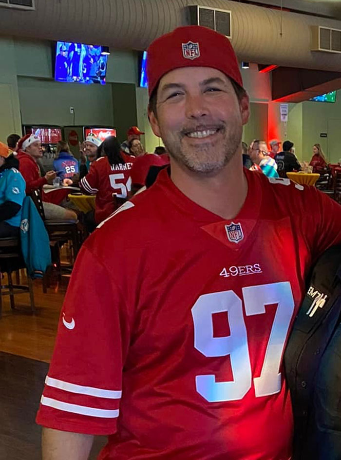 Man in a red NFL 49ers jersey and cap smiling in a crowded sports bar during a game day gathering.