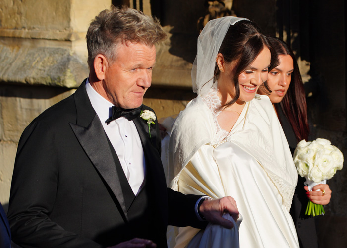 Gordon Ramsay with his daughter wearing a satin bed sheet wedding dress, smiling and walking outdoors in sunlight.