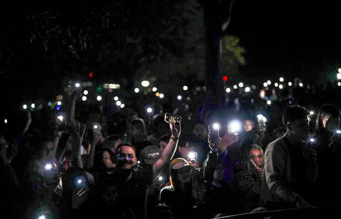 Crowd holding up phones with lights on at night, illustrating public space reserved on New Year’s Eve slammed by locals. Crowd holding up phones with lights on at night, illustrating public space reserved on New Year’s Eve slammed by locals.