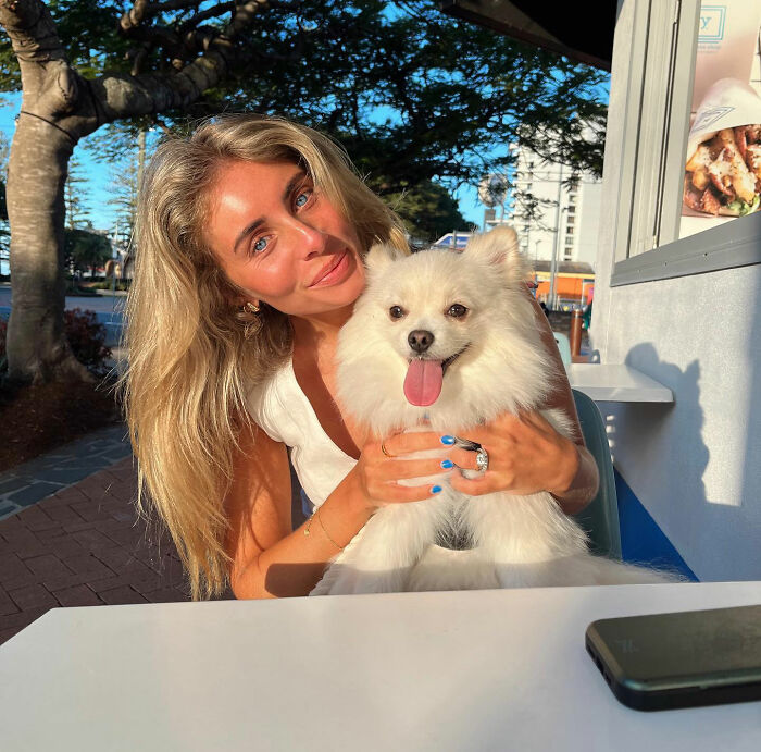 Blonde woman holding a fluffy white dog outdoors in bright sunlight, related to Bonnie Blue controversy in Bali.
