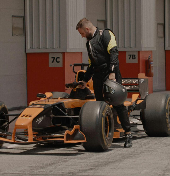Man in racing suit holding helmet beside orange and black race car in pit area, unrelated to crypto scammer details. Man in racing suit holding helmet beside orange and black race car in pit area, unrelated to crypto scammer details.