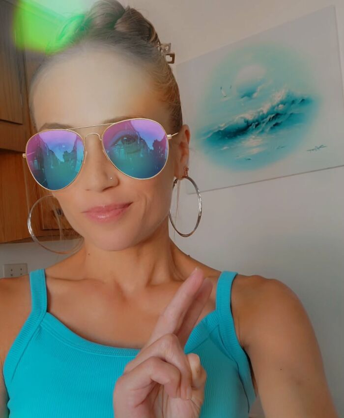 Woman wearing reflective sunglasses and hoop earrings, posing indoors with fingers crossed, related to racist Cinnabon employee story. Woman wearing reflective sunglasses and hoop earrings, posing indoors with fingers crossed, related to racist Cinnabon employee story.