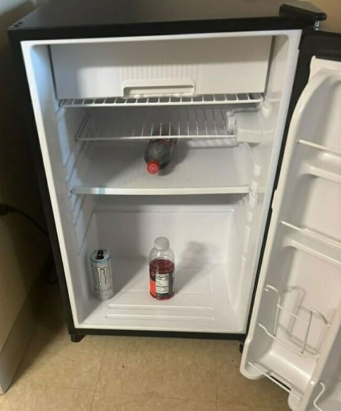 Small dude fridge with sparse contents includes a soda bottle, energy drink can, and sports drink on empty shelves.