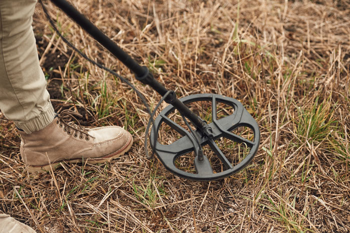 Person wearing tan boots using a metal detector on dry grass in the woods searching for hidden objects.