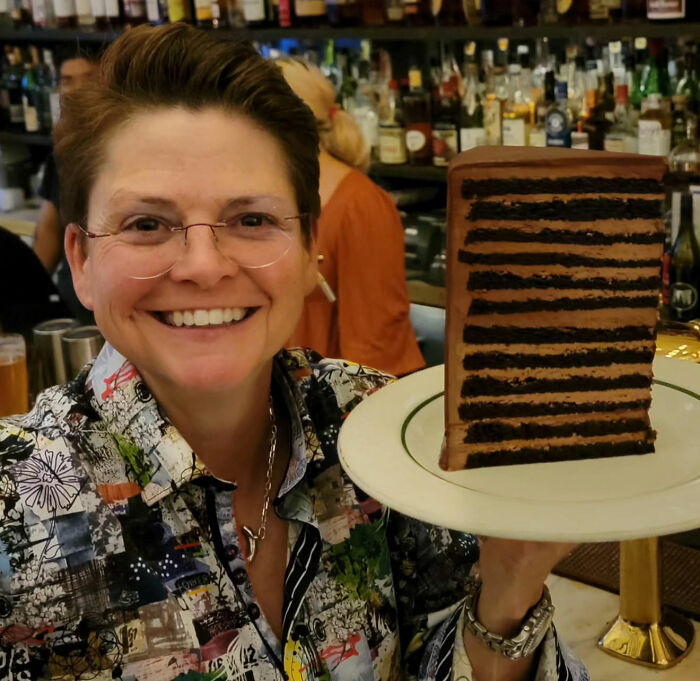 Smiling woman at a bar holding a plate with a large slice of layered chocolate cake, NYC setting.