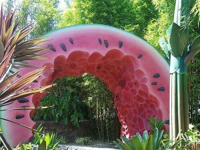 Archway shaped like a giant watermelon slice surrounded by lush plants creating a trippy picture from another reality.