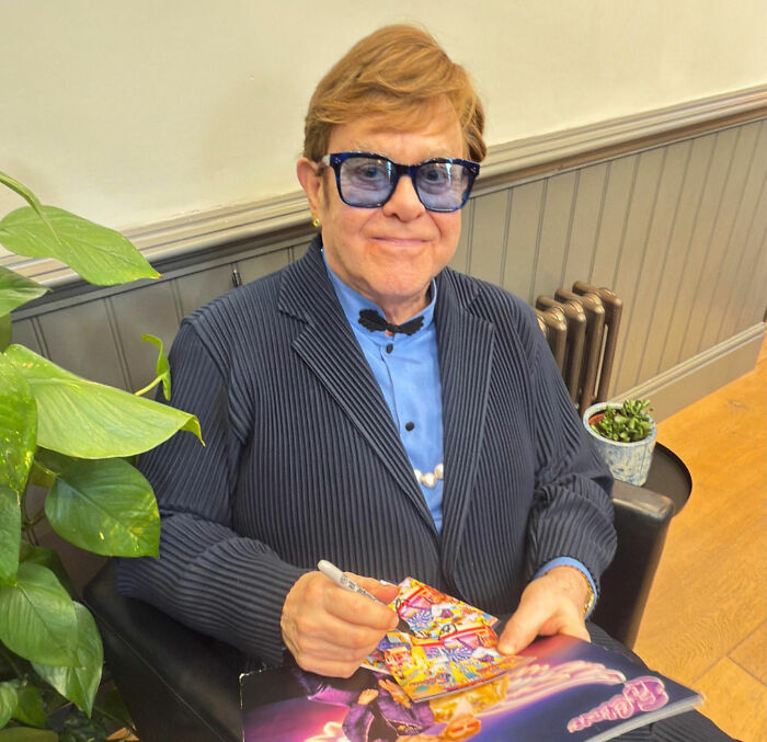 Elton John wearing blue glasses and pinstripe suit, sitting indoors signing colorful album cover with plants nearby Elton John wearing blue glasses and pinstripe suit, sitting indoors signing colorful album cover with plants nearby