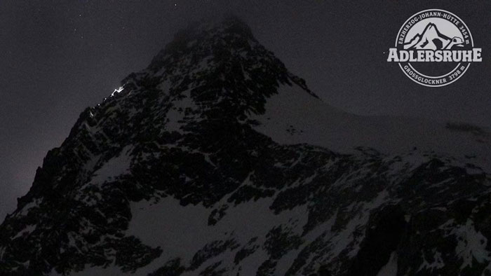 Snowy peak of Austria&rsquo;s highest mountain at night with climbers' lights and Adlersruhe emblem in the sky corner.