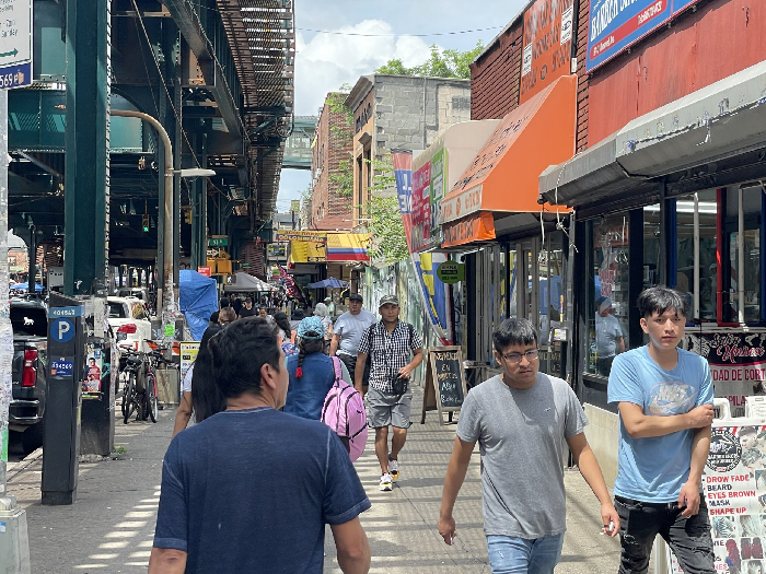 Busy New York City street scene under elevated train tracks with diverse pedestrians and colorful storefronts.