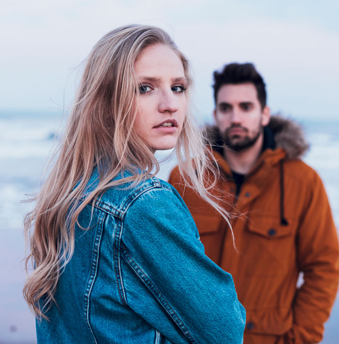 Young woman in denim jacket and man in brown coat at the beach illustrating things women do to seem attractive men can’t stand