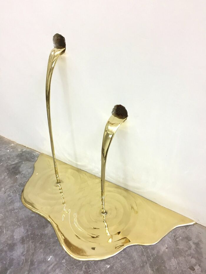 Abstract golden sculpture with liquid-like streams pouring from wall mounts onto a reflective surface in a trippy picture.