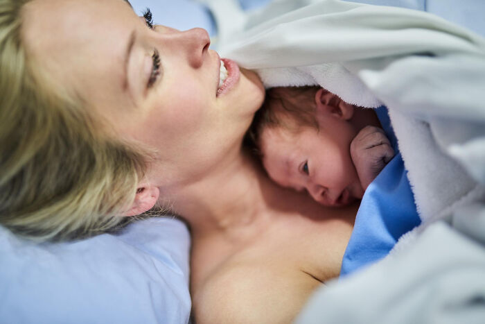Mother holding newborn baby skin to skin after delivery in a hospital setting, while men missed delivery moments nearby.