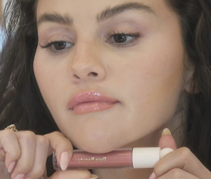 Selena Gomez close-up holding lip gloss, responding to fan questions about how she shaves her mustache.