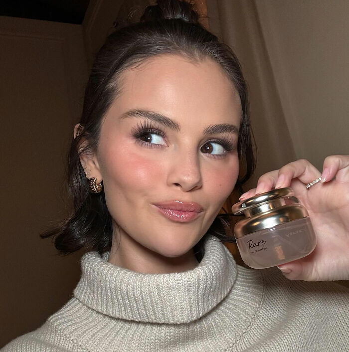 Selena Gomez holding Rare beauty product, smiling and looking to the side with flawless makeup and styled hair indoors.