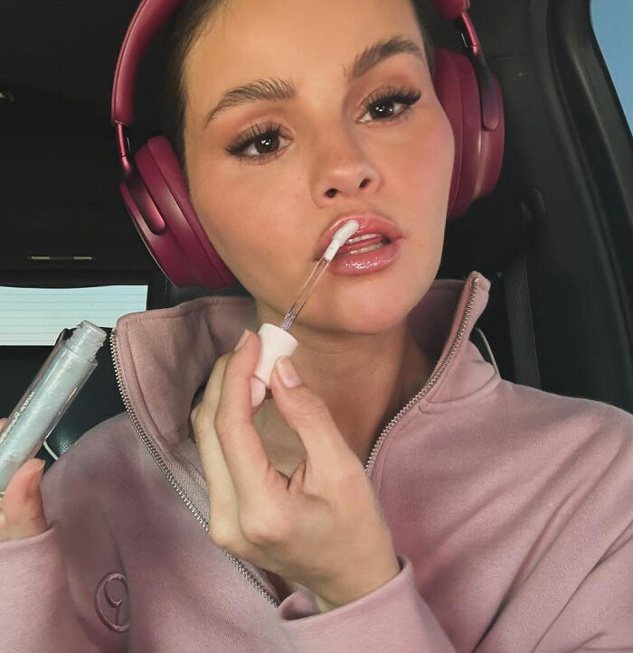 Selena Gomez applying lip gloss wearing headphones in a car, related to fan question about how she shaves her mustache.