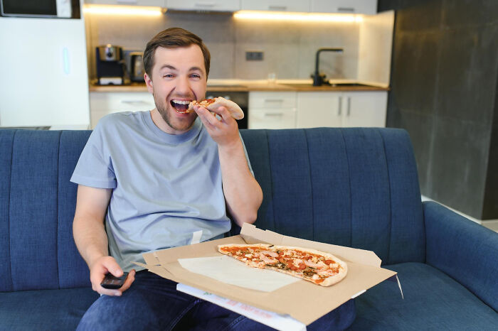 Man eating pizza on couch holding remote, illustrating men missed delivery babies concept in casual home setting.