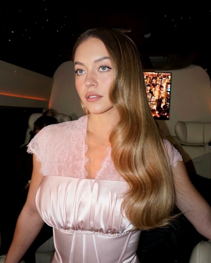 Sydney Sweeney with long wavy hair and soft makeup in a pink satin dress, highlighting her viral MAGA makeover look.