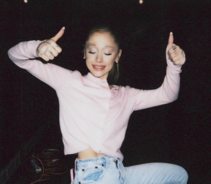 Ariana Grande wearing a light pink sweater and jeans, giving thumbs up with eyes closed against a black background.