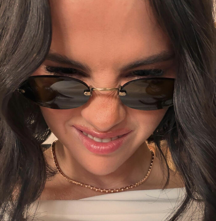 Selena Gomez smiling, wearing sunglasses and a gold chain necklace in a close-up photo.