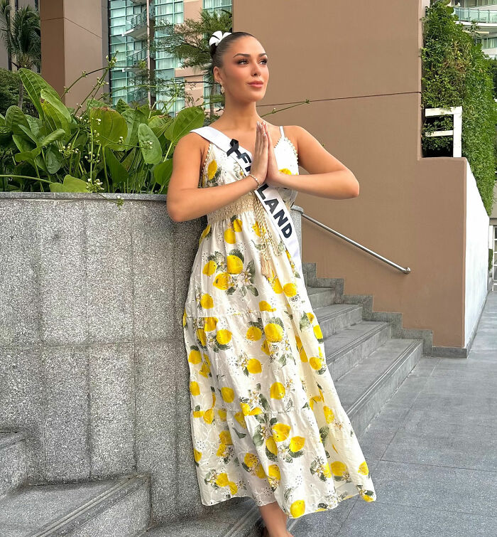 Beauty queen from Finland in a floral dress posing with hands in a prayer gesture near stairs outside a building.