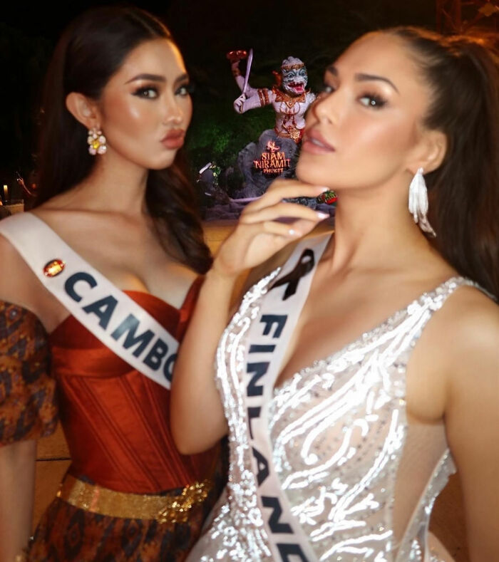 Two beauty queens wearing Cambodia and Finland sashes posing at night with a statue in the background.