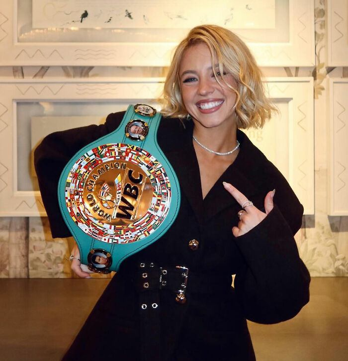 Sydney Sweeney smiling and holding a colorful WBC championship belt indoors in a casual black outfit.