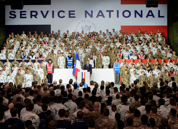 French soldiers standing behind President Macron during a national service event, highlighting odd behavior and abrupt stage exit.