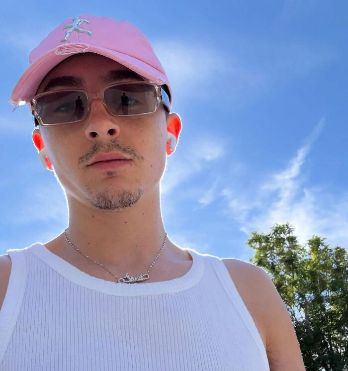 Young man wearing a pink cap and sunglasses, posing outdoors under a clear blue sky, showing a casual red carpet look.