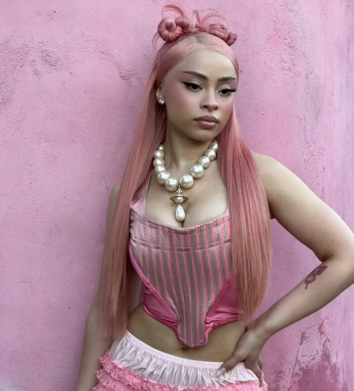 Ice Spice posing against a pink wall wearing a pink corset and pearl necklace sparking outfit outrage among fans.