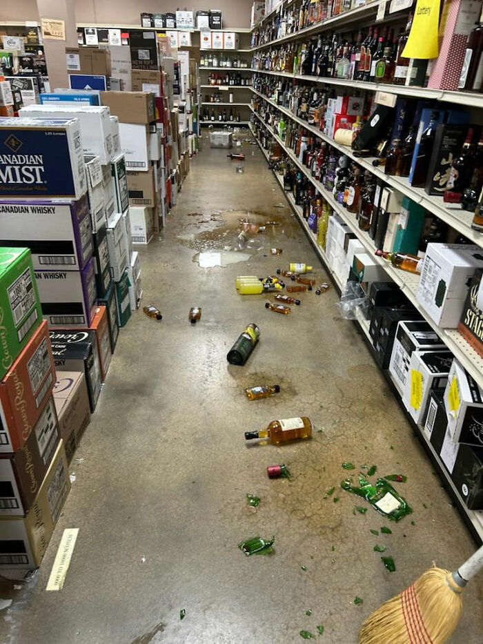 Liquor store aisle with broken bottles and spilled alcohol after a raccoon raid causes chaotic mess on the floor.
