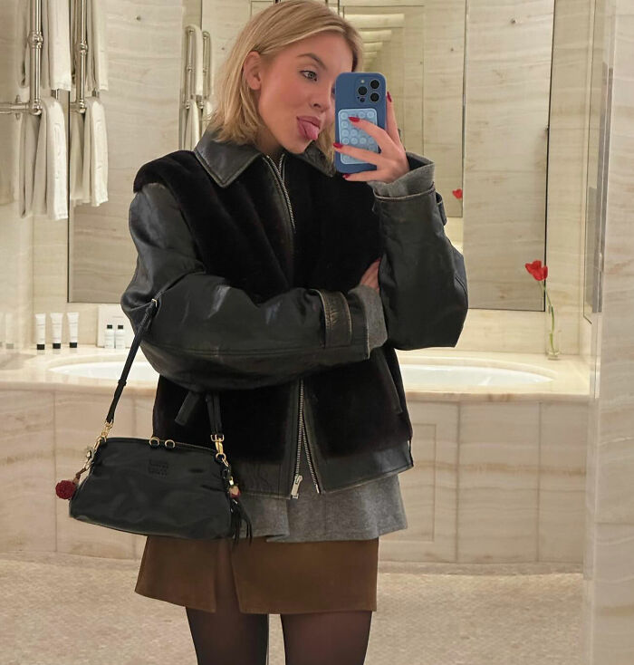 Sydney Sweeney taking a mirror selfie indoors, wearing a black leather jacket and holding a small black handbag.