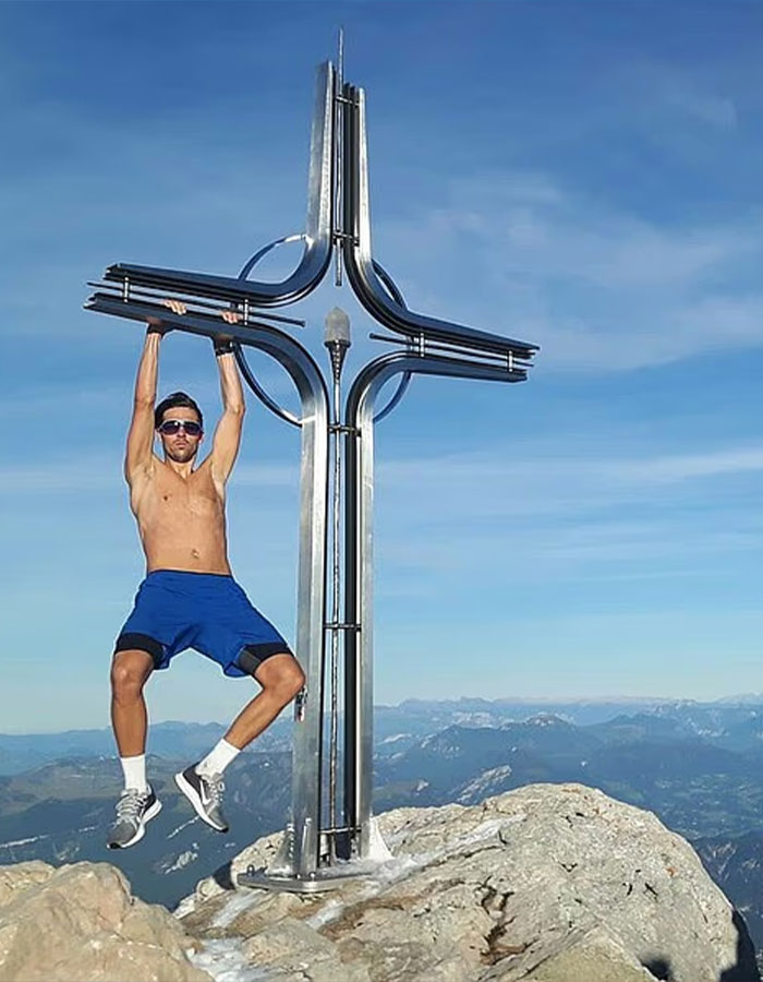 Man wearing sunglasses and blue shorts hanging from metal cross on Austria&rsquo;s highest peak with mountain landscape behind him