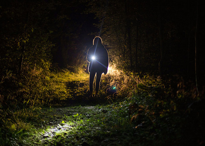 Person holding flashlight walking on a dark forest path at night, evoking creepy and mysterious remote isolated places.