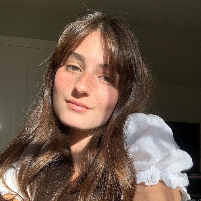 Young woman with long brown hair and freckles, softly smiling in natural sunlight, unrelated to Rob Reiner&rsquo;s son arrest news.