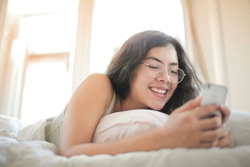 Young woman smiling while reading a good morning message to my love on her smartphone in a cozy bedroom setting Young woman smiling while reading a good morning message to my love on her smartphone in a cozy bedroom setting