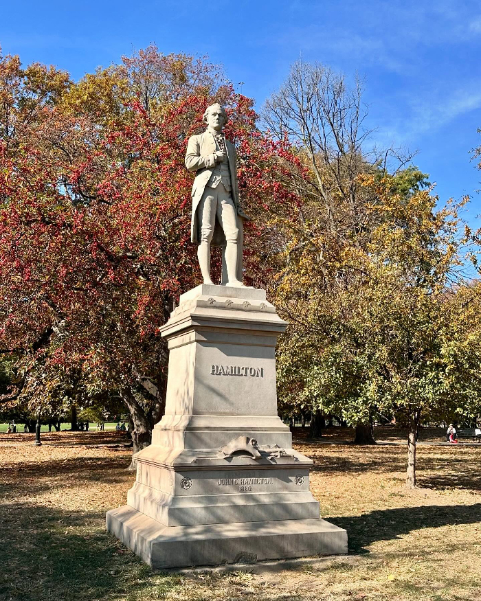 Statue of Alexander Hamilton in a New York City park surrounded by autumn trees under a clear blue sky.