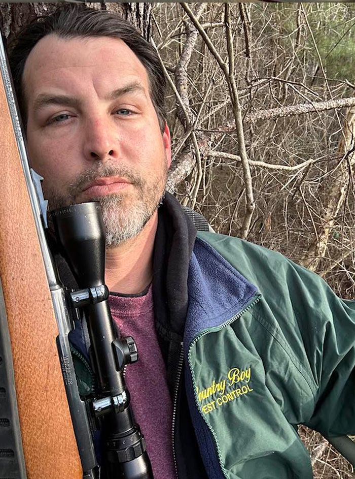 Man outdoors holding rifle with scope, wearing green jacket, amid bare tree branches in a tense NFL argument context.