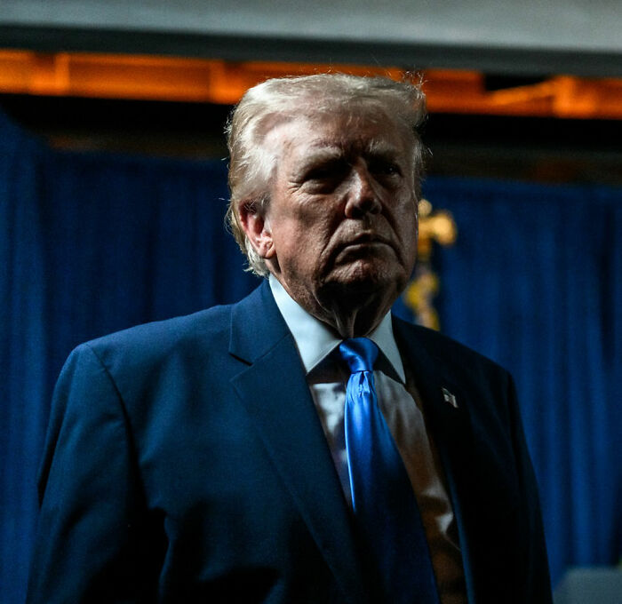Donald Trump in a dark suit and blue tie, standing in front of blue curtains during a dimly lit event.
