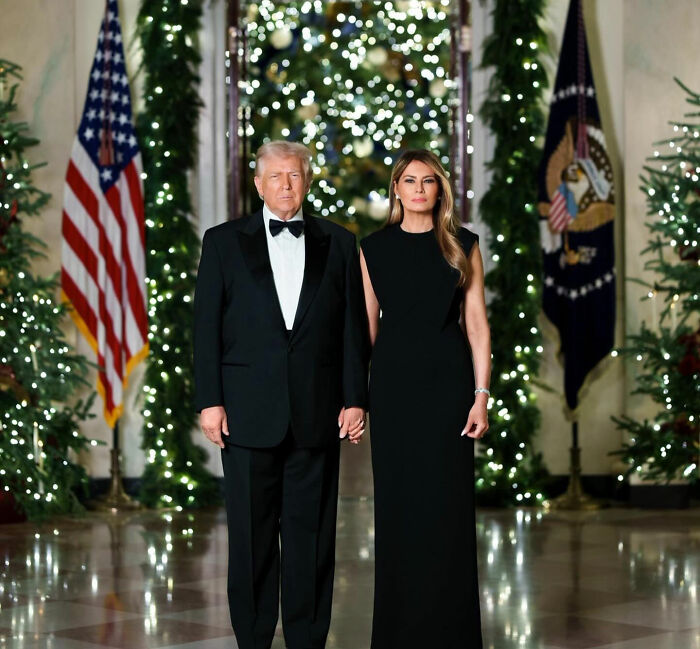 Donald Trump and Melania Trump in formal attire standing by Christmas trees with festive lights during holiday event.