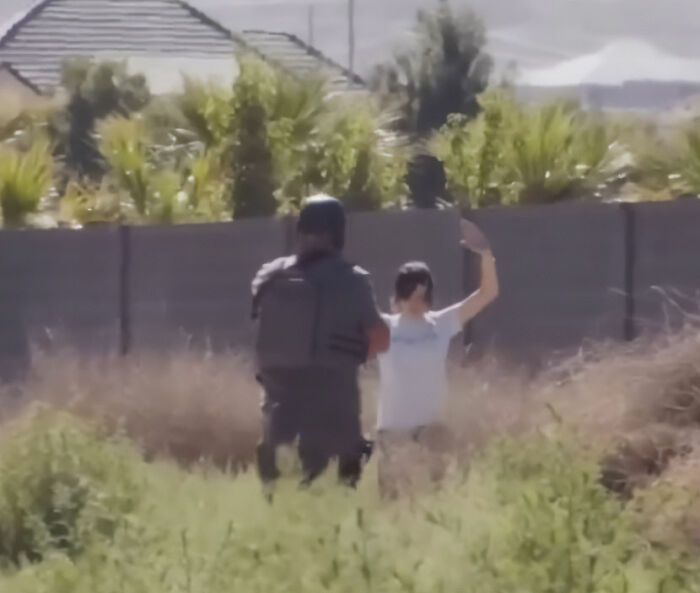 Trans Muslim woman raising hands while facing armed officer outside near a fence in an open grassy area. Trans Muslim woman raising hands while facing armed officer outside near a fence in an open grassy area.